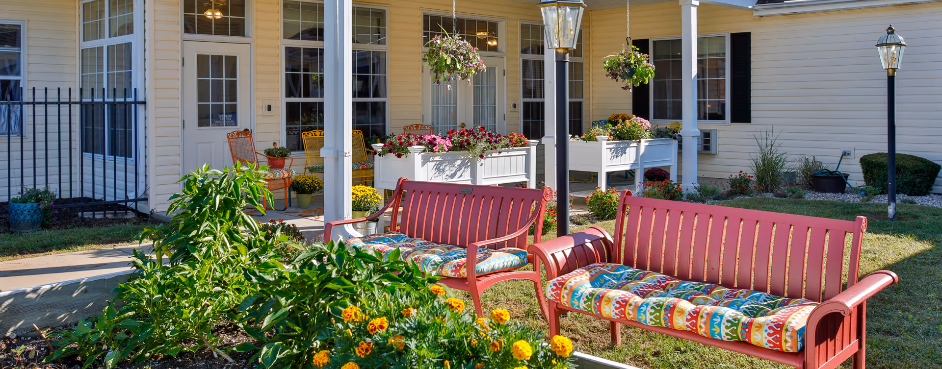 Outdoor patio area at 1019 Belle's Place of Crawfordsville featuring two red benches with colorful cushions, hanging flower baskets, potted plants, and a garden with various flowers. The patio is adjacent to a building with cream-colored siding and multiple windows and doors.