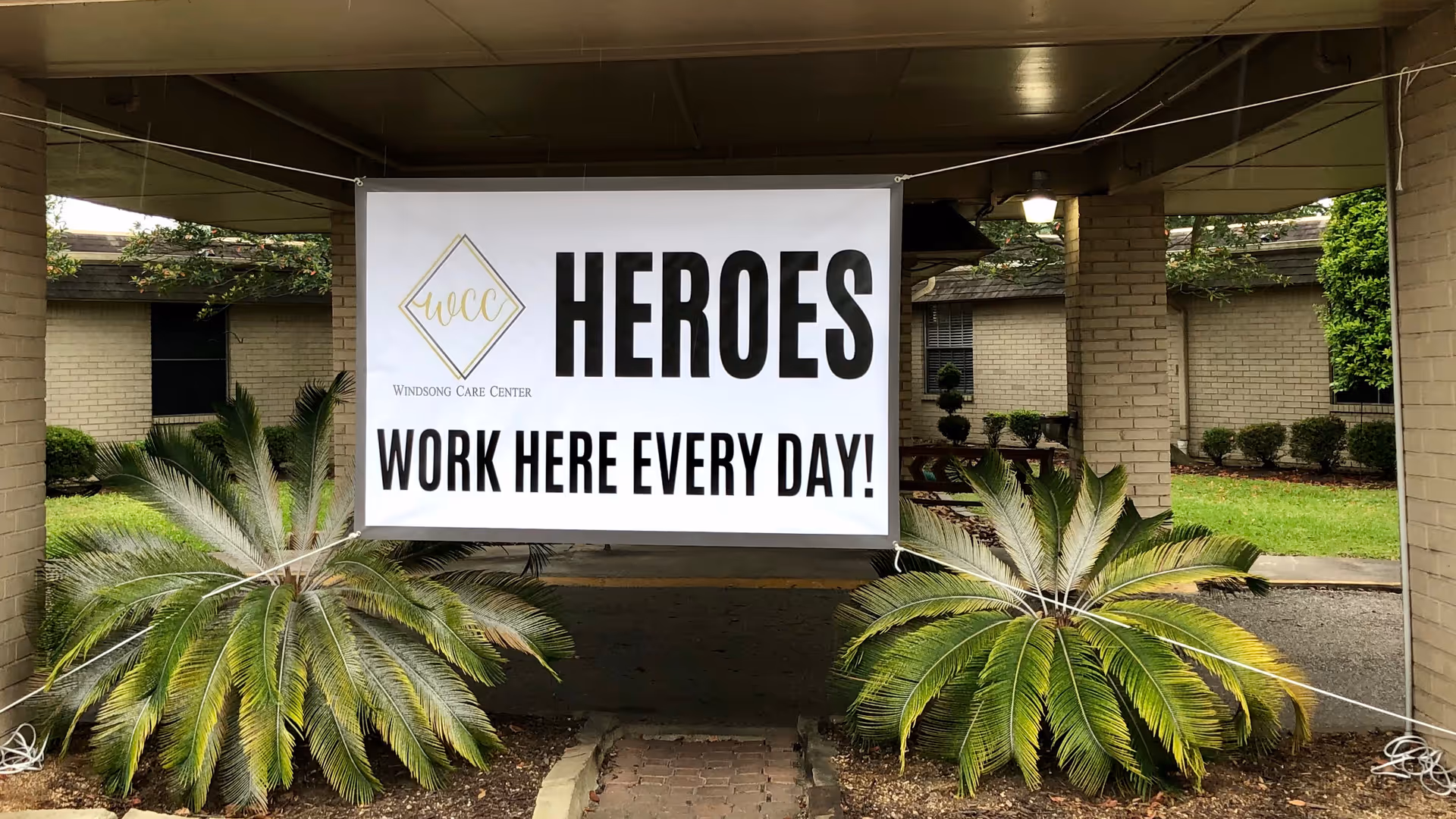 Covered entrance with a large banner reading "HEROES WORK HERE EVERY DAY!" hung between brick columns above landscaped plants.
