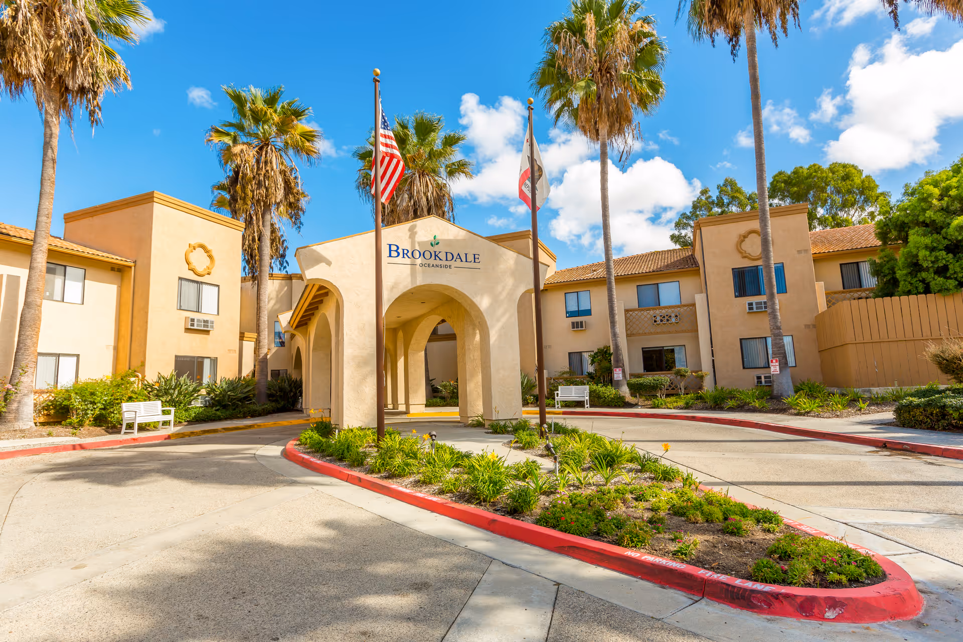 Sunny exterior view of a Brookdale Senior Living building with palm trees and flags.