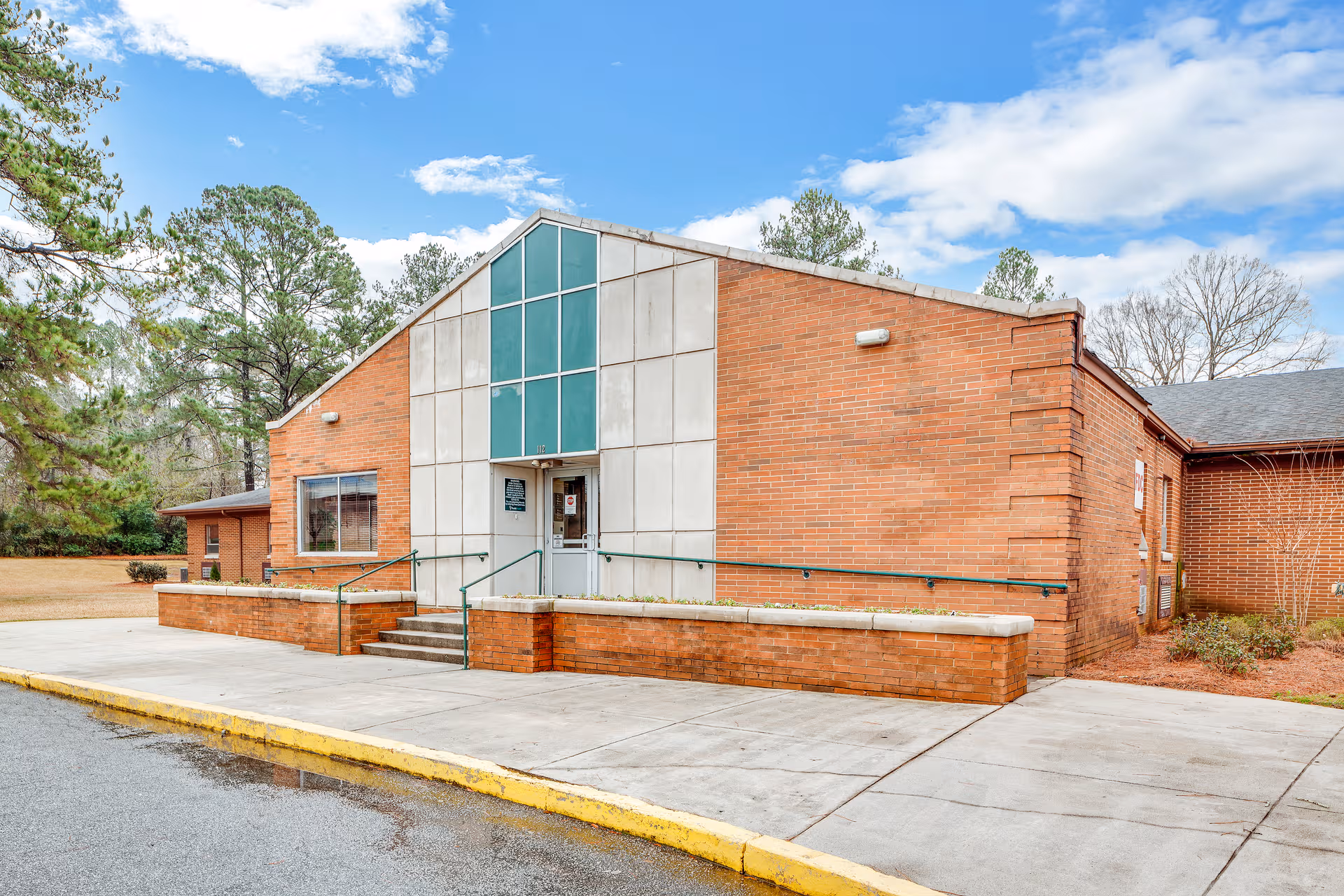 Brick senior living facility exterior with glass entrance, ramp, and surrounding trees.
