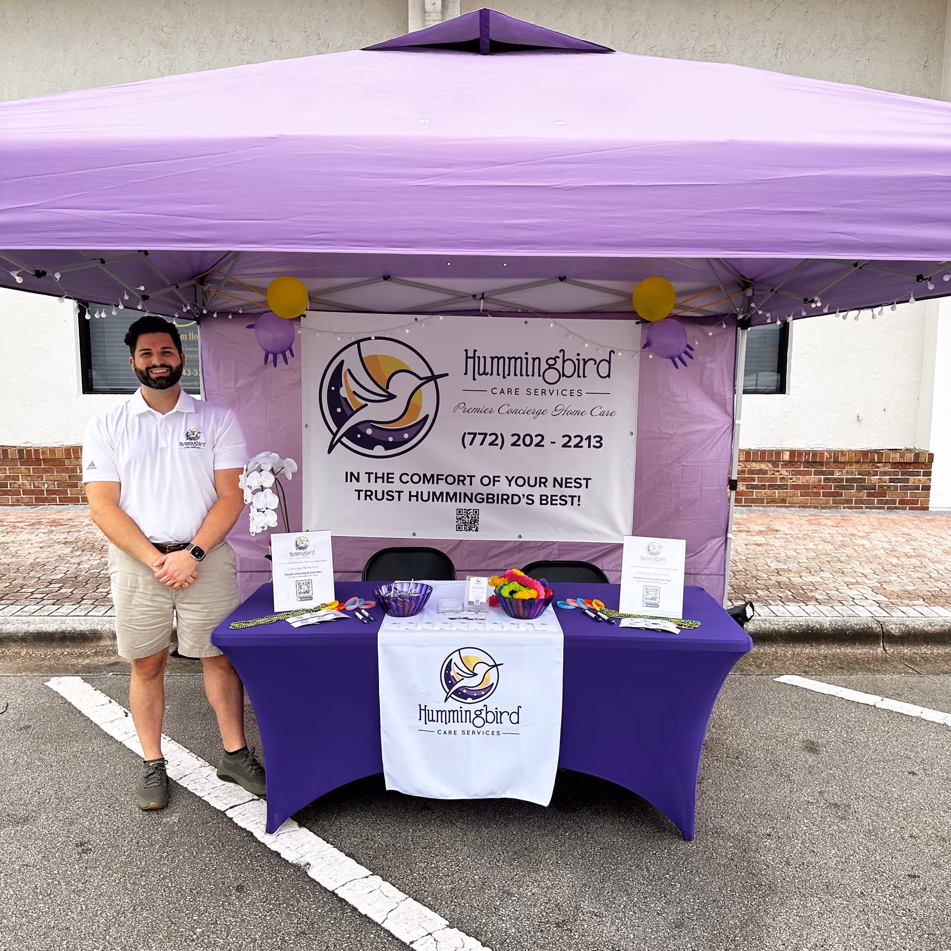 A man stands beside a purple tent booth displaying the Hummingbird Care Services banner.
