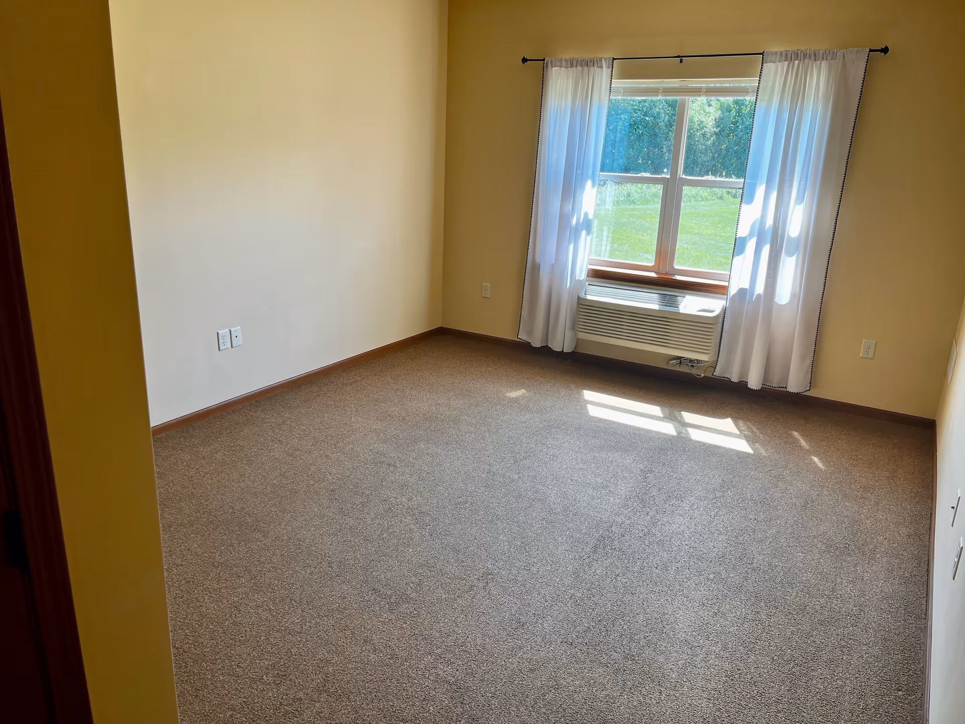 Empty room with beige walls and carpeted floor, a window with white curtains letting in sunlight, and an air conditioning unit below the window.