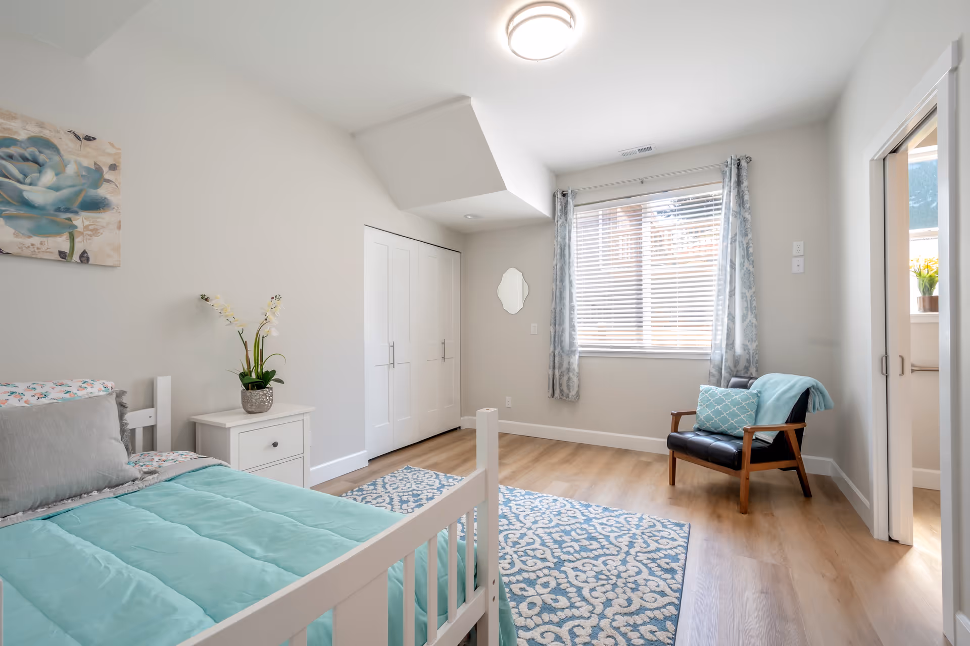 Bright bedroom with a white bed, nightstand, chair, window, and closet in soft decor.