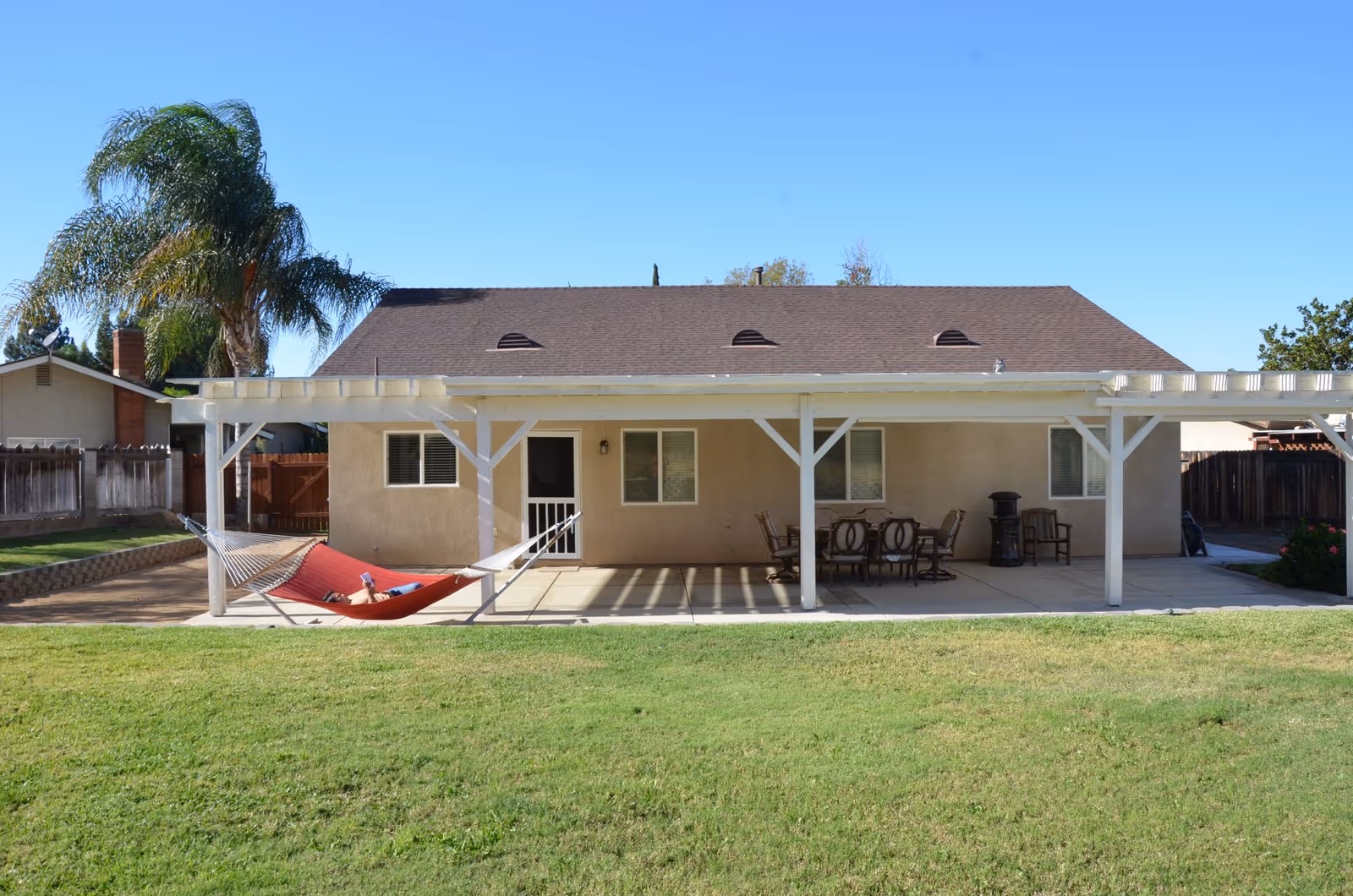 Backyard view of a single-story house with a brown roof and beige walls. There is a covered patio with white wooden beams, a red hammock with a person lying on it, and an outdoor dining table with chairs. The yard has green grass and a palm tree on the left side.