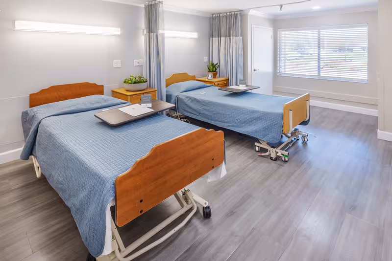 A bright and clean room with two hospital-style beds covered with blue bedspreads. Each bed has a wooden headboard and footboard, and a small overbed table. There are two wooden nightstands between the beds, each with a plant and some items on top. The room has light gray walls, large windows with blinds, and light wood flooring.