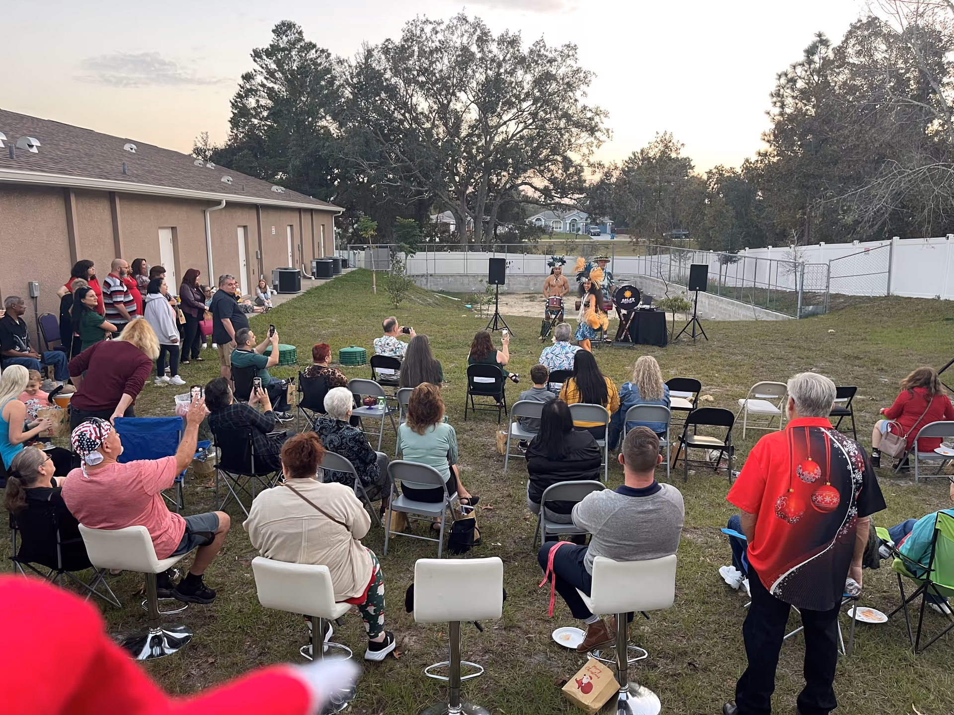 Outdoor gathering of residents seated in chairs watching a small performance outdoors.