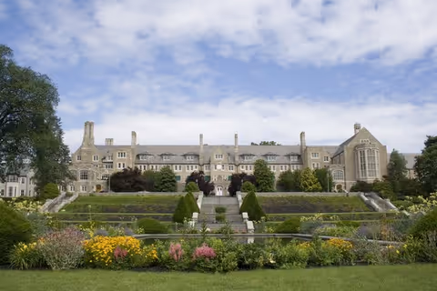 Large stone institutional building with terraced formal gardens, colorful flower beds and a lawn under a partly cloudy sky.