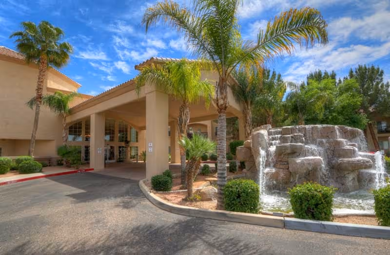 Exterior view of The Montecito Senior Living facility entrance with a covered driveway, palm trees, and a rock waterfall feature under a partly cloudy blue sky.