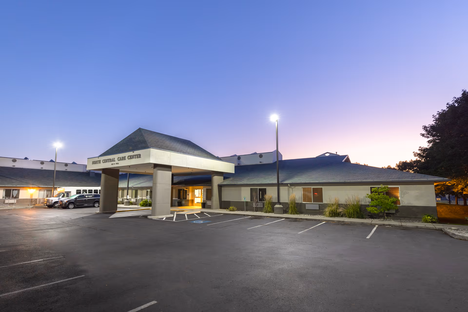 Front entrance of the North Central Care Center building at dusk with a covered drive-through, parked cars, and an empty parking lot.