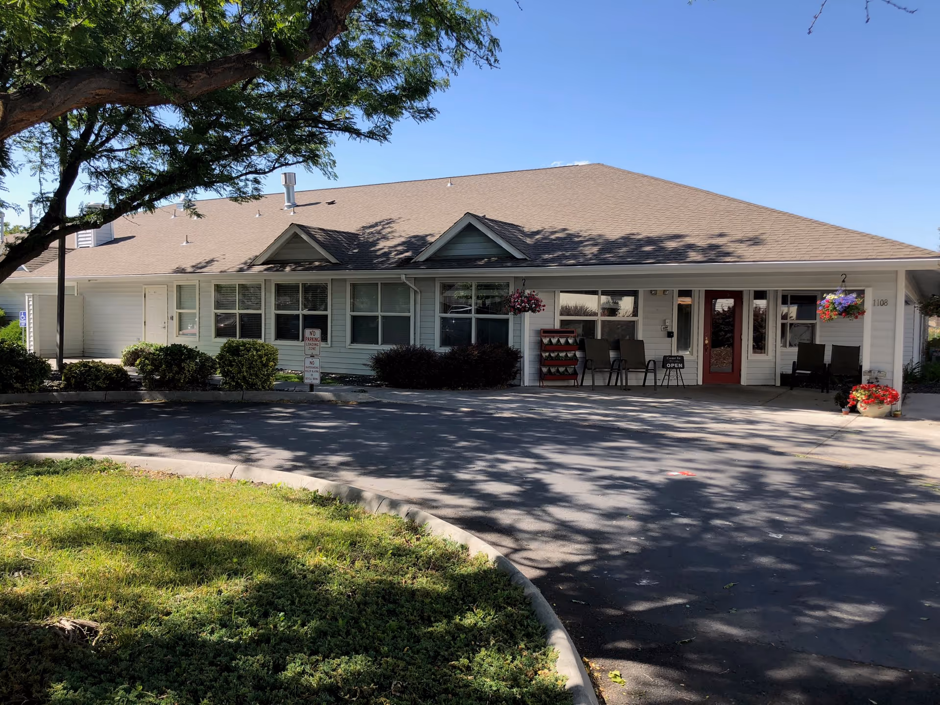Single-story assisted living facility front with a covered entrance, chairs, and hanging flower baskets.