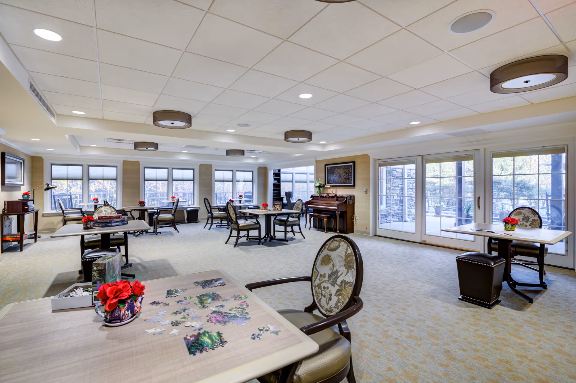 Bright senior living common area with round tables, chairs, windows, and a piano.