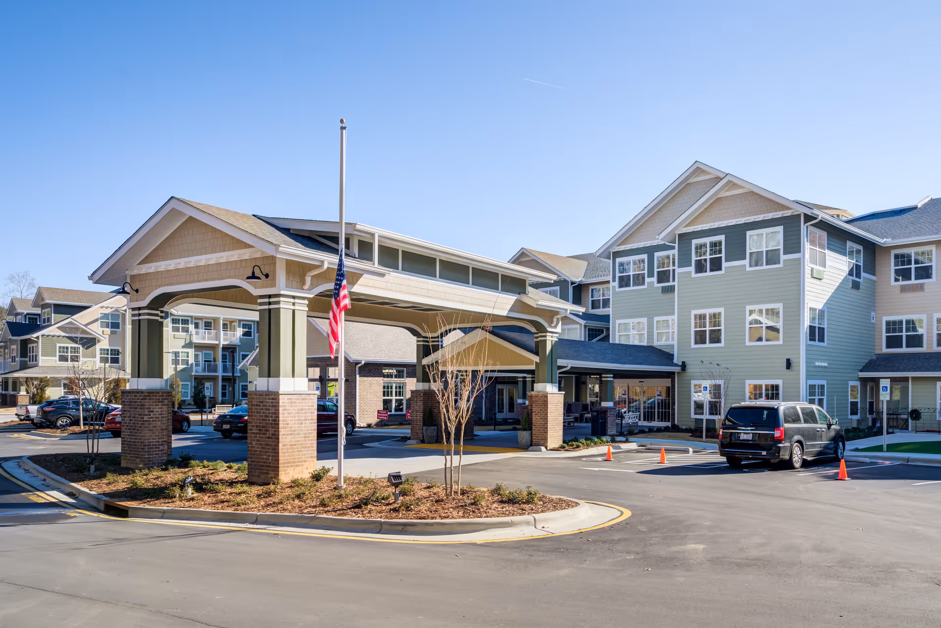 Front entrance and porte-cochère of a multi-story senior living building with an American flag and parked cars.
