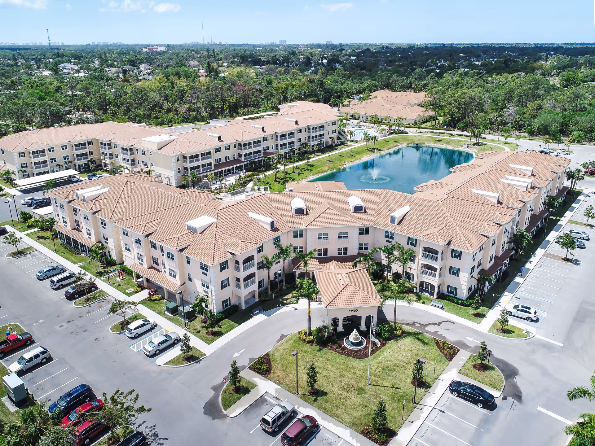 Aerial view of a senior living complex with terracotta-roofed buildings surrounding a central pond and fountain.