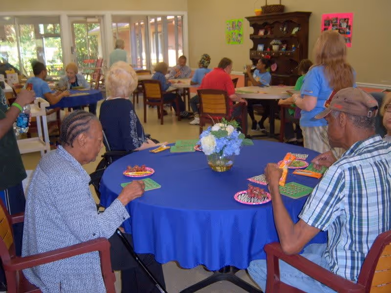 Several elderly individuals seated around tables with blue tablecloths in a bright room, engaging in activities such as playing bingo. The room has large windows letting in natural light and a wooden cabinet against the wall with decorations. A vase with flowers is on the table in the foreground.