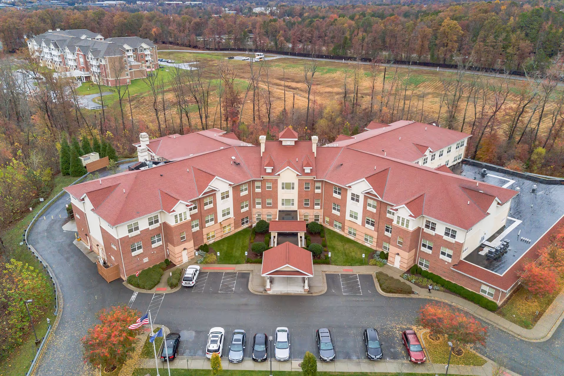 Aerial view of a large senior living facility with a red roof and multiple wings forming a U-shape. The building is surrounded by trees with autumn foliage and has a parking lot with several cars in front. There is a covered entrance at the center of the building.