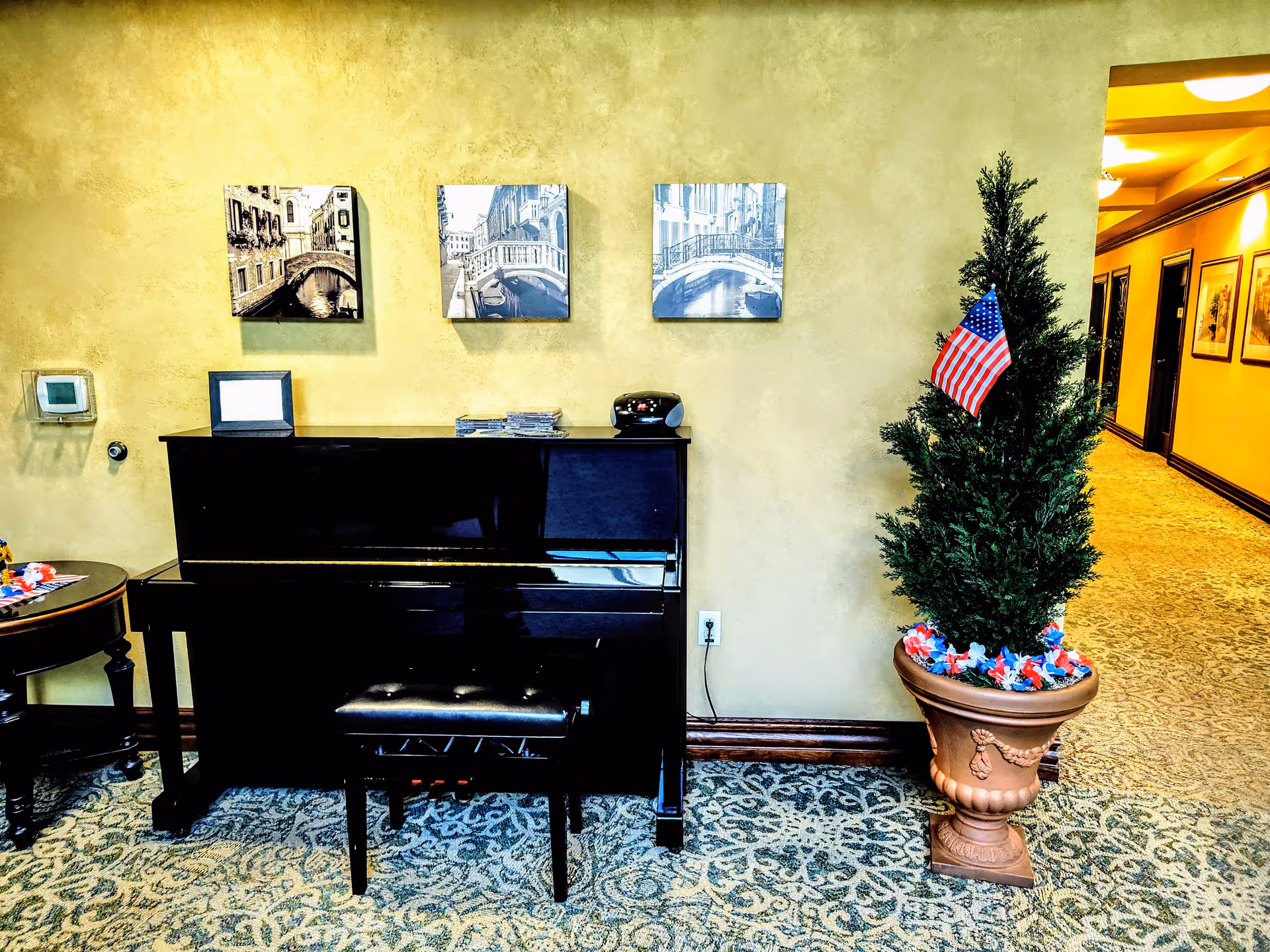 Black upright piano beside beige wall, framed photos, round table, and a potted Christmas tree.