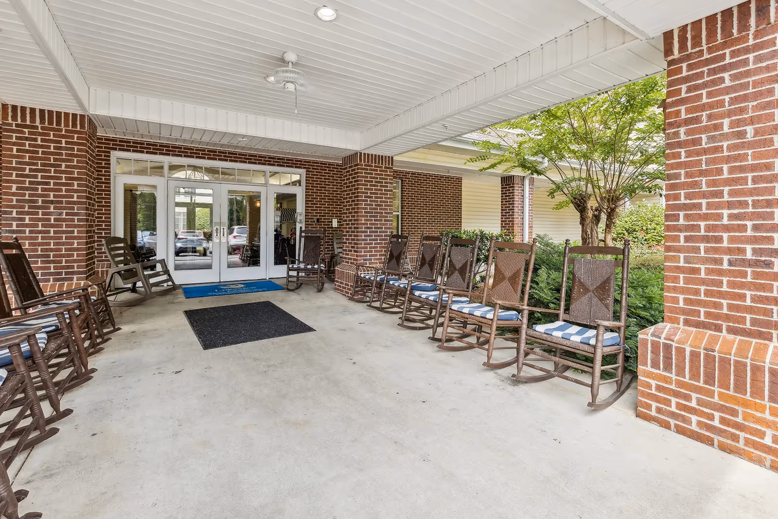 Covered entrance area of a building with brick walls and multiple wooden rocking chairs with blue and white striped cushions arranged along the sides. There are double glass doors leading inside, a ceiling fan, and some greenery visible on the right side.