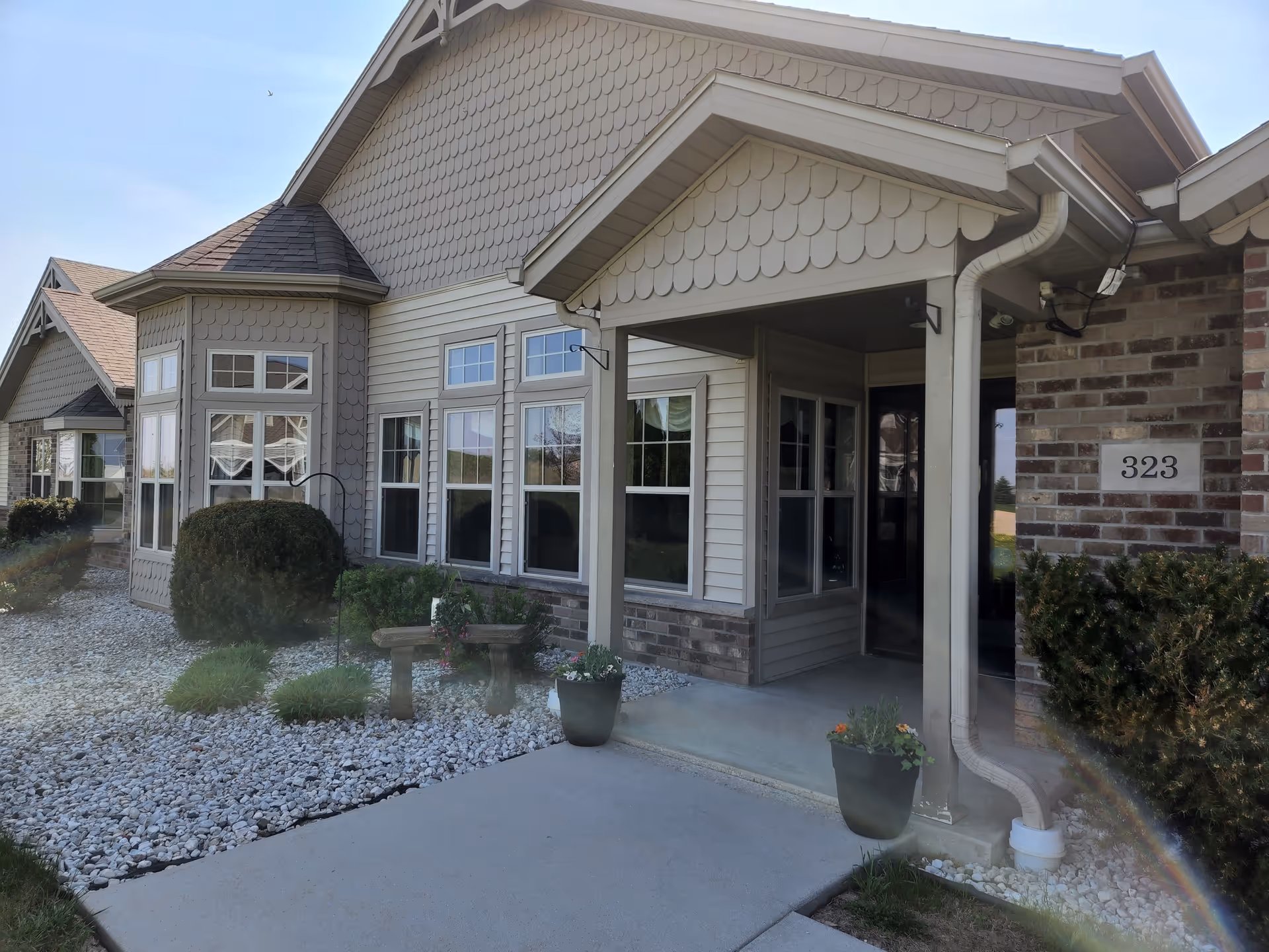 Exterior view of ABridge Care Cottage of Chilton showing the entrance with a covered porch, beige siding, brick accents, multiple windows, and landscaping with bushes and potted plants.