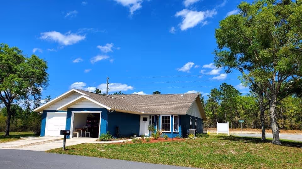 Single-story blue house with a gray roof under a bright blue sky with scattered clouds. The house has a garage with the door open, a white front door, and several windows. There are trees and grass surrounding the house, and a paved road in front.