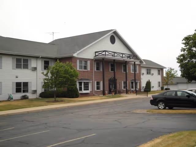 Exterior front view of a two-story senior living building with a brick central facade, white siding wings, entrance portico, and a parking lot with a car.
