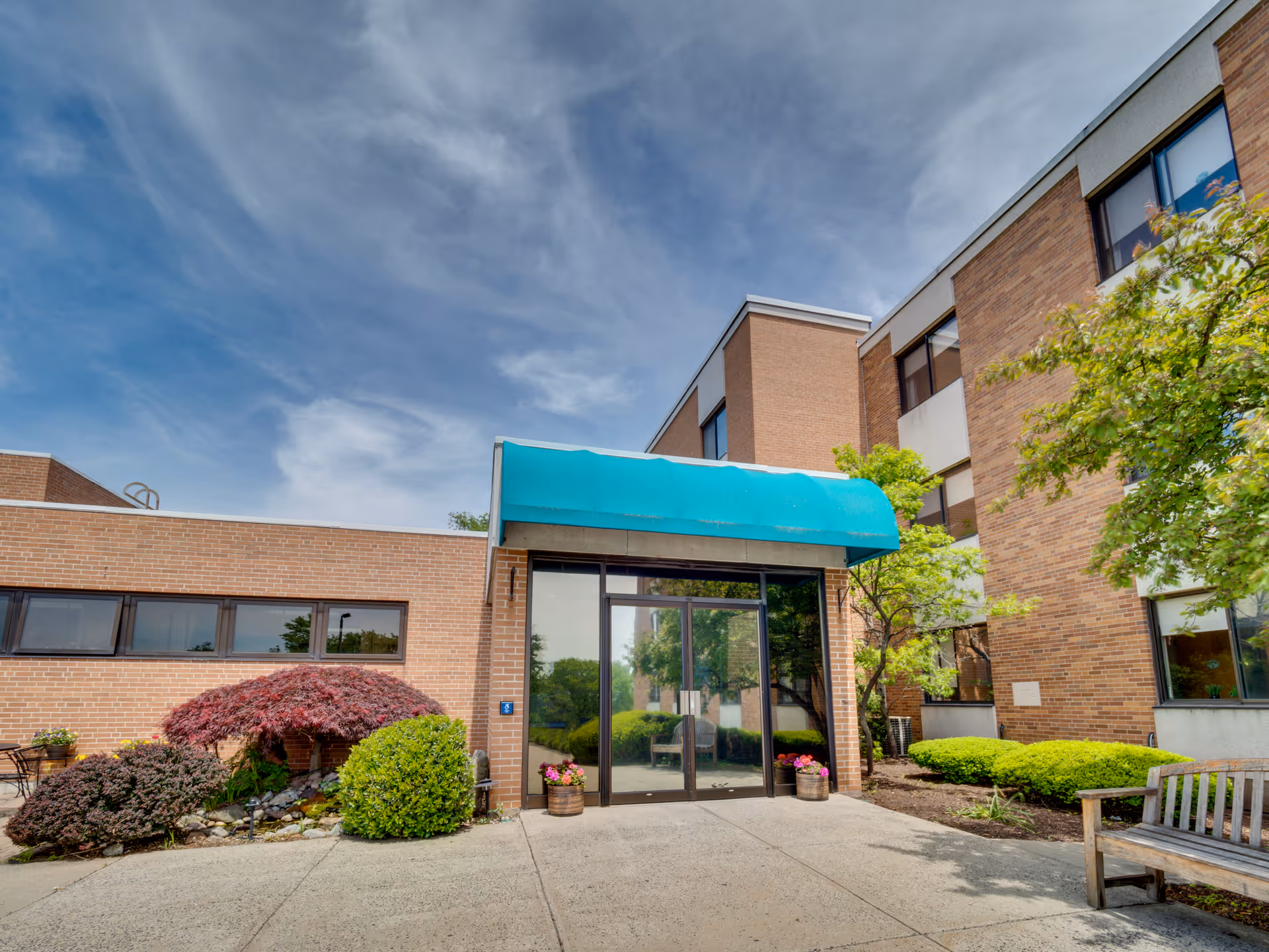 Brick senior living facility entrance with teal awning and glass doors surrounded by landscaping.