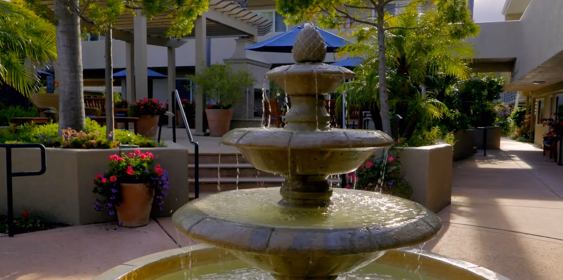 Tiered stone fountain in a sunny landscaped outdoor courtyard with potted flowers, trees, and seating areas.