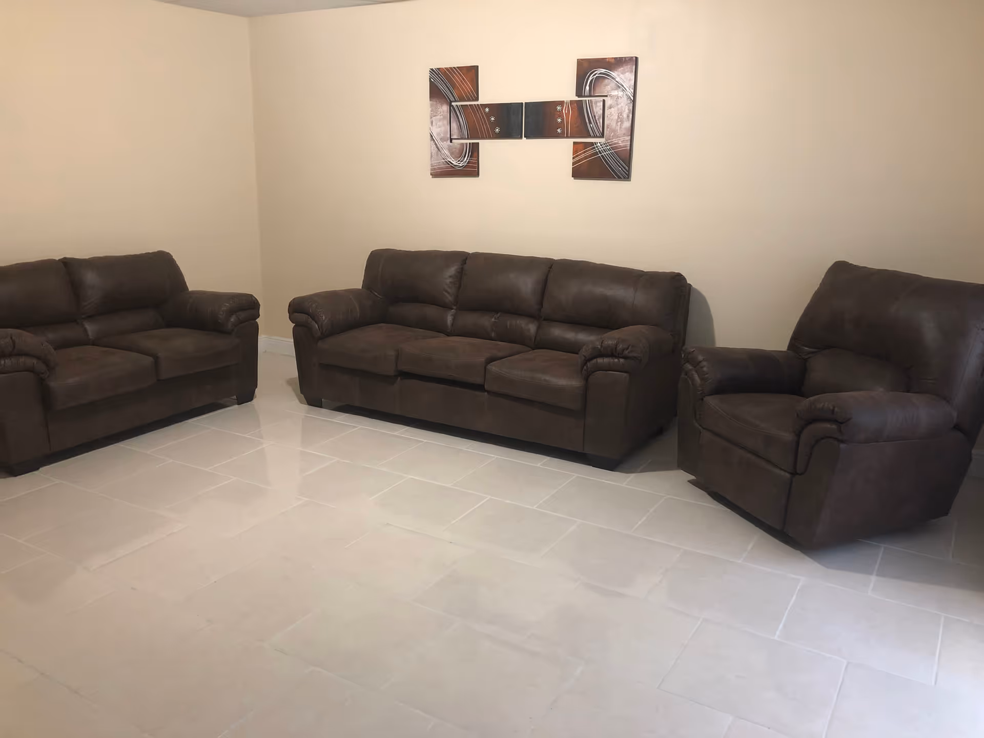 Three brown leather sofas arranged in a spacious, tiled living room with abstract wall art.