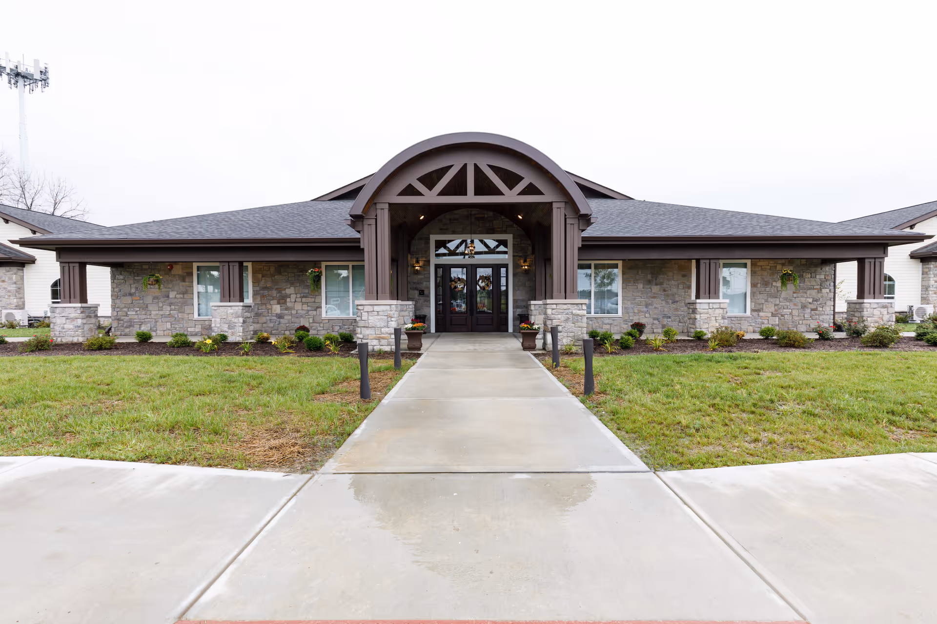 Front exterior view of a single-story building with stone and wood accents, a covered entrance with an archway, and a concrete walkway leading to double glass doors. The surrounding area has green grass and small shrubs.