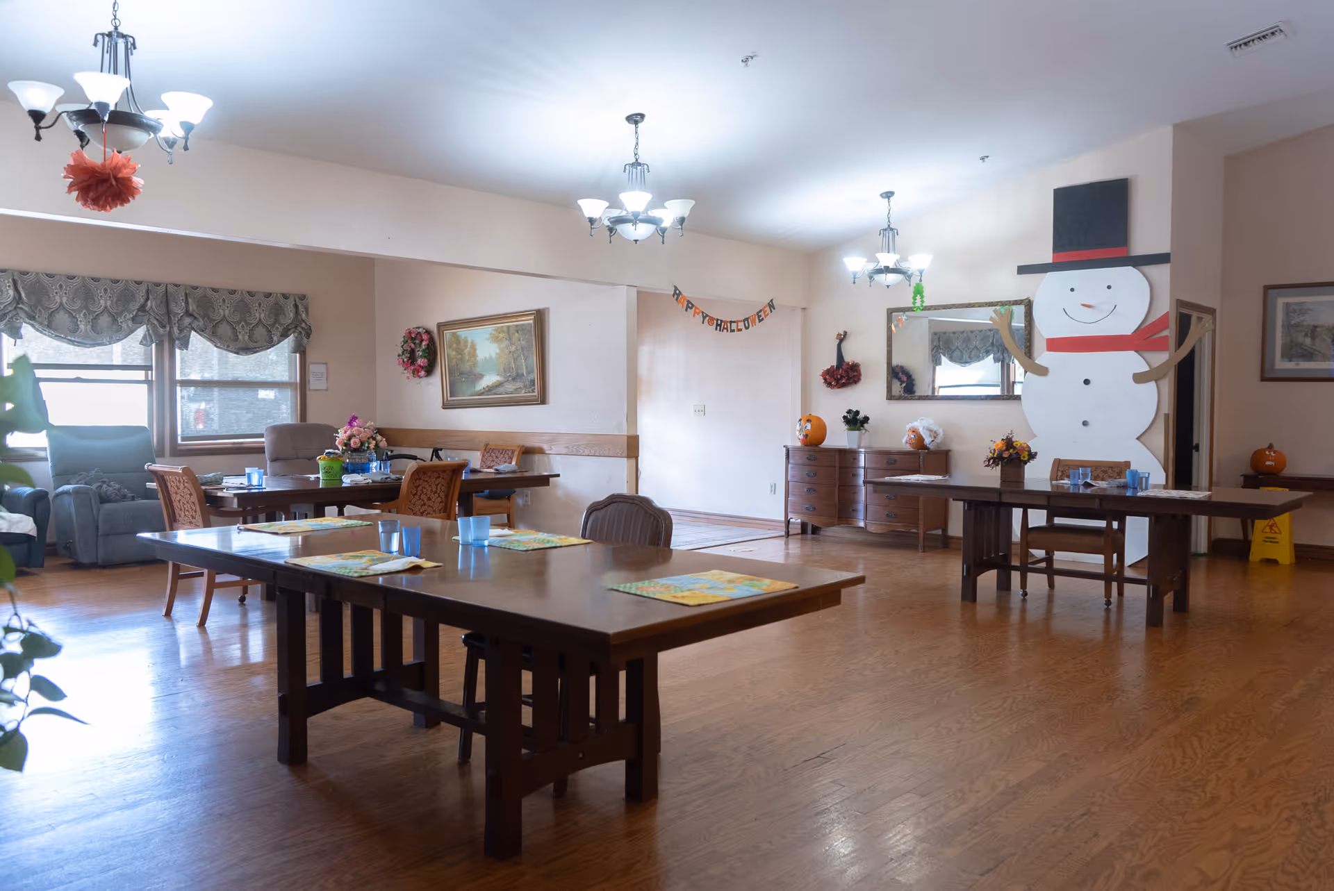 Spacious senior living dining area with large wooden tables, chairs, and festive snowman decor.