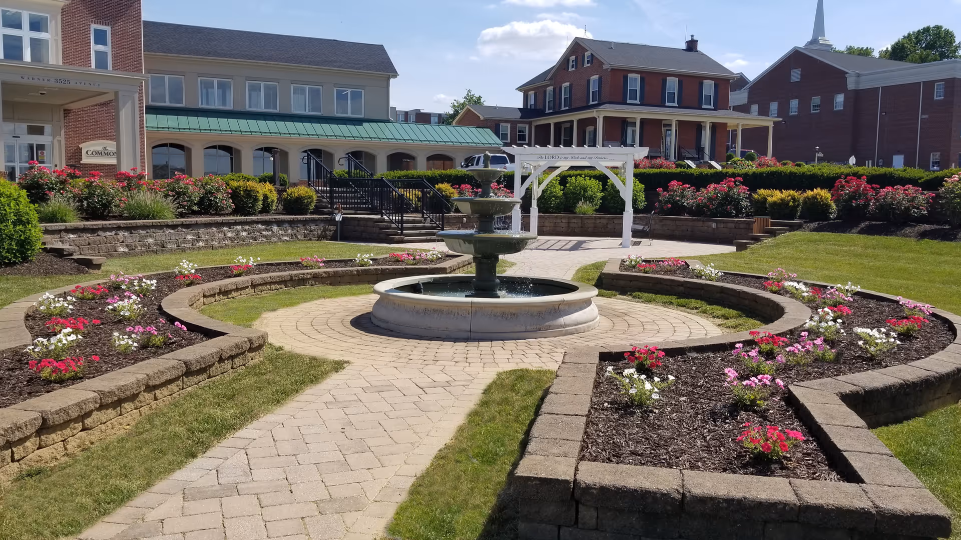 Landscaped courtyard with a central fountain, paved walkways, flower beds, and retirement community buildings in the background.