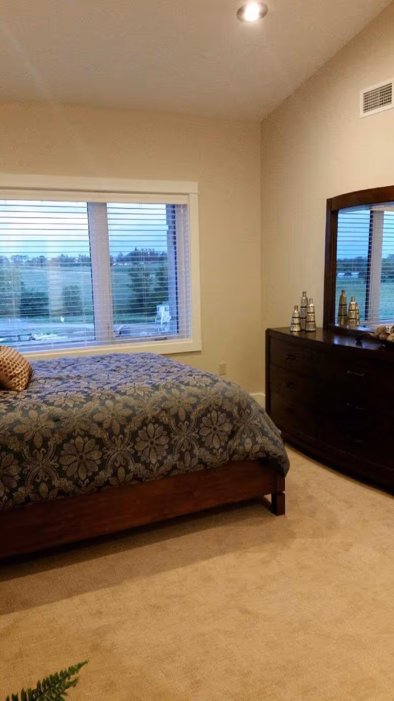 A well-lit bedroom with a patterned bed, large window with blinds, and a dark wood dresser with mirror.