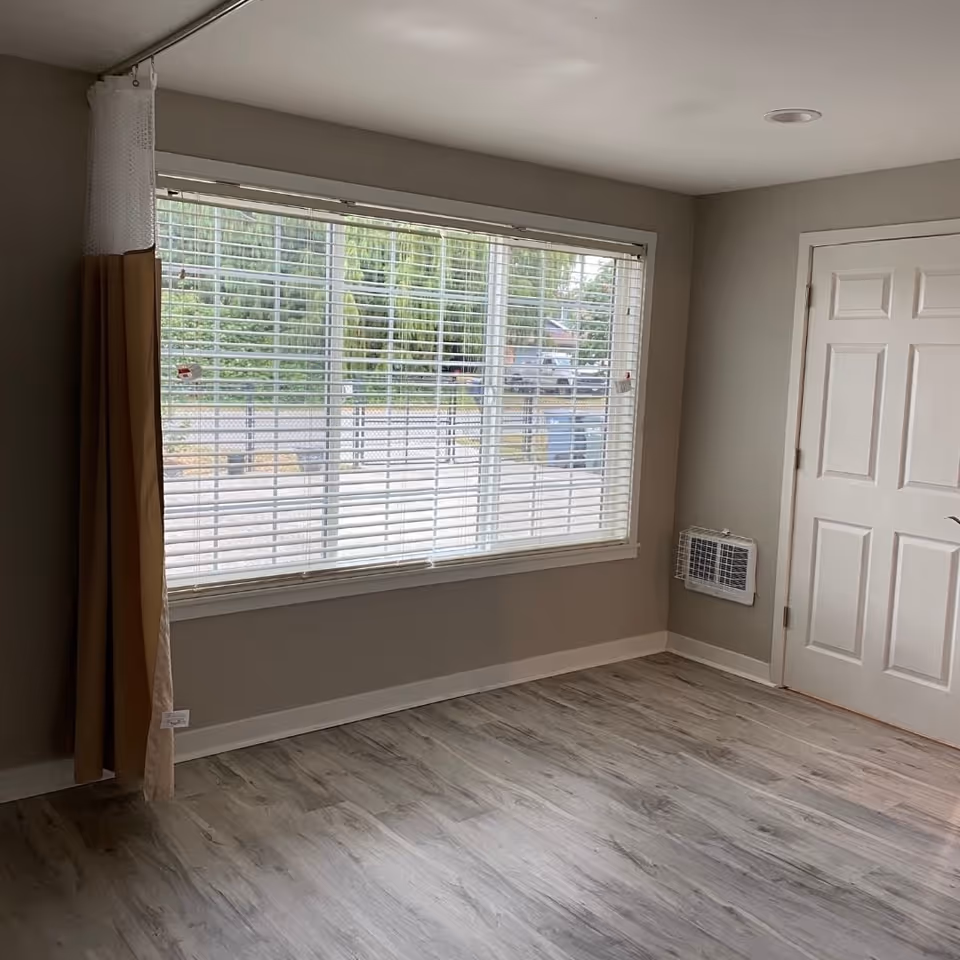 Empty, gray-toned room with large window blinds, light wood flooring, and a white door.