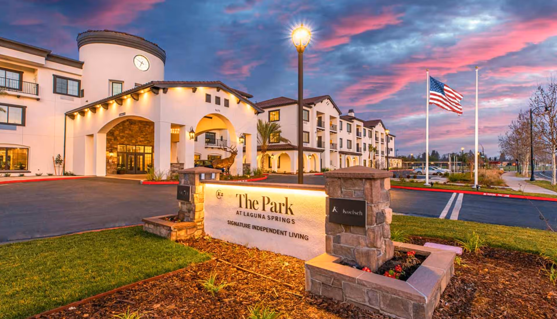 Exterior front entrance of The Park at Laguna Springs senior living building with an illuminated sign, flagpoles, and a colorful sunset sky.