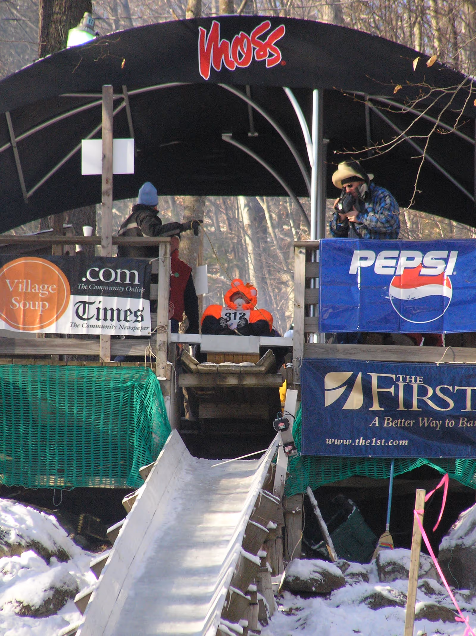 Outdoor wooden platform with banners, people in winter clothing, and a snowy ramp.