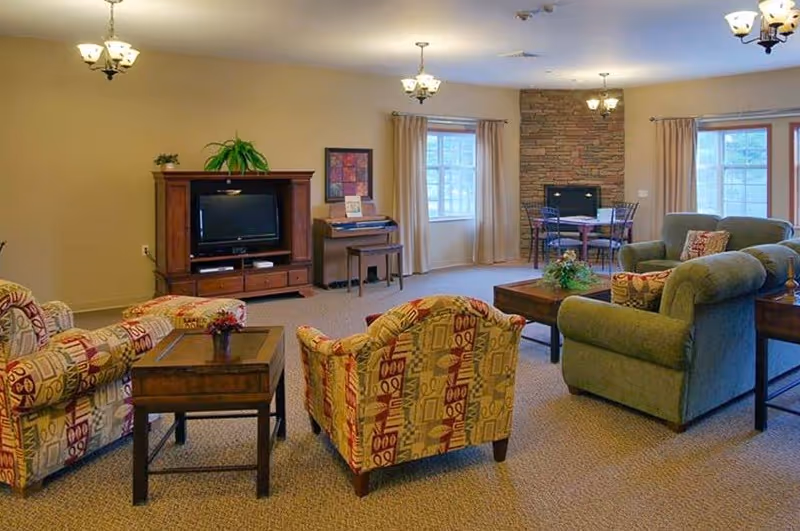 Spacious furnished common living room with patterned armchairs, a green sofa, TV cabinet, coffee tables and a small dining area by windows.