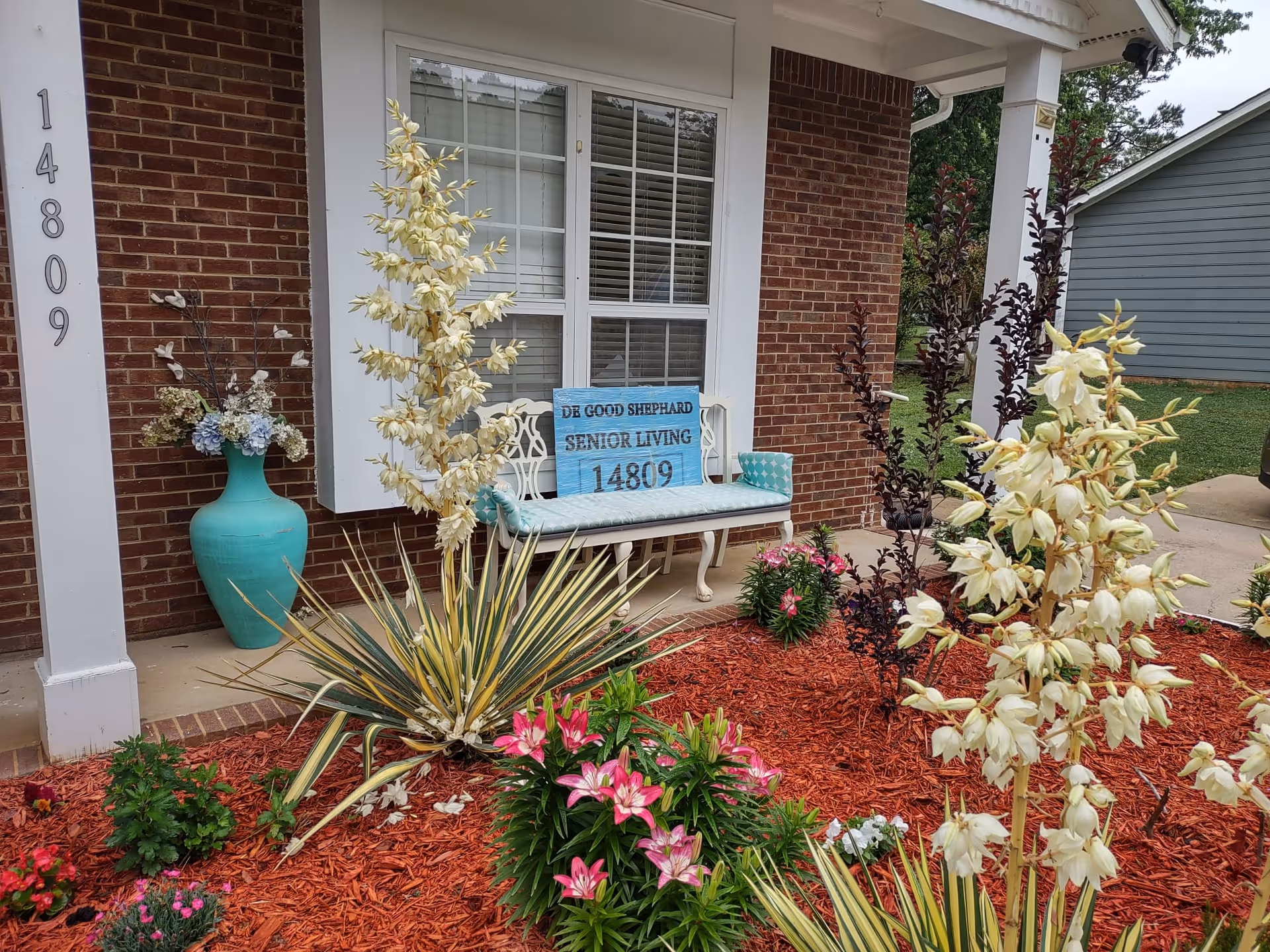 Front porch of a brick senior living building with a bench, a blue sign reading 'DE GOOD SHEPHARD SENIOR LIVING 14809', potted decor and a landscaped flowerbed.