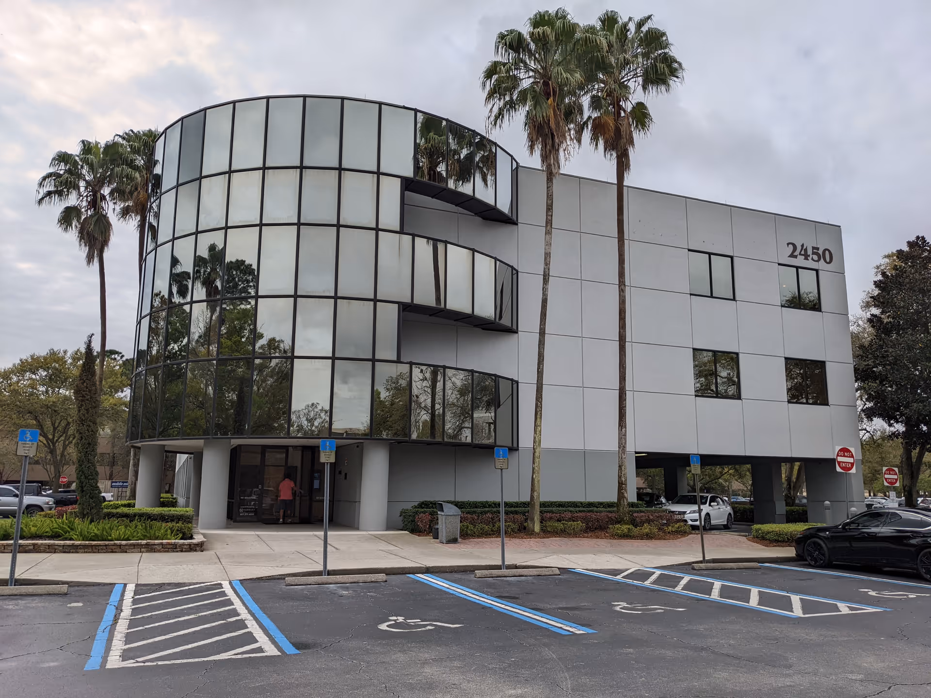 Modern curved glass-front building with palm trees and a parking lot.