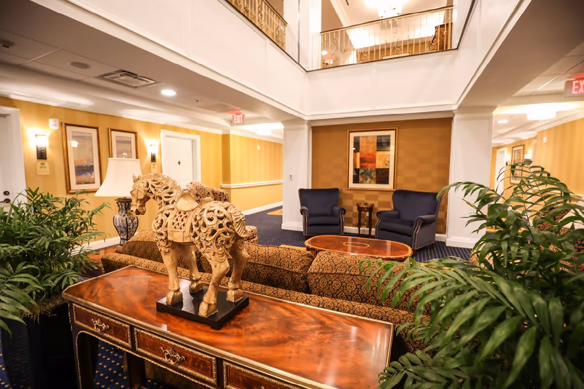 A well-lit interior common area with a decorative wooden horse sculpture on a polished wooden table in the foreground. The room features patterned sofas, two blue armchairs, a wooden coffee table, framed artwork on the walls, potted plants, and a balcony railing above.