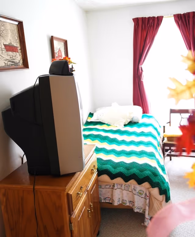 A bedroom with a twin bed, crocheted blanket, dresser, and a bulky TV facing the bed.