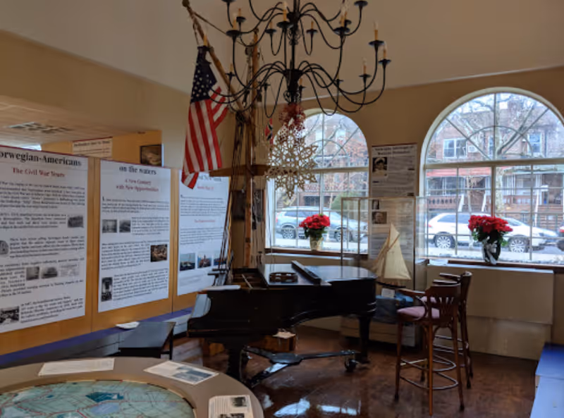 Common room interior with a grand piano under a chandelier, informational display panels, an American flag, arched windows and poinsettias.