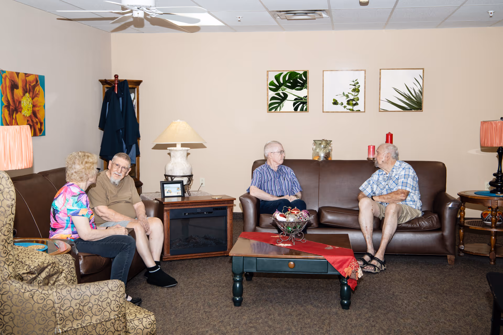 A cozy living room in a retirement community with four elderly people sitting and conversing on two brown leather sofas. The room features a coffee table with a red runner and decorative centerpiece, a side table with a lamp, wall art with botanical prints, and a ceiling fan.