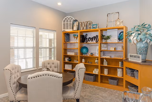 A cozy interior room with a round wooden table surrounded by four beige upholstered chairs with nailhead trim. Behind the table is a large wooden shelving unit decorated with various home decor items including vases, framed pictures, and decorative signs. Two windows with white blinds allow natural light into the room. A large blue vase with a leafy plant sits on a side cabinet to the right.