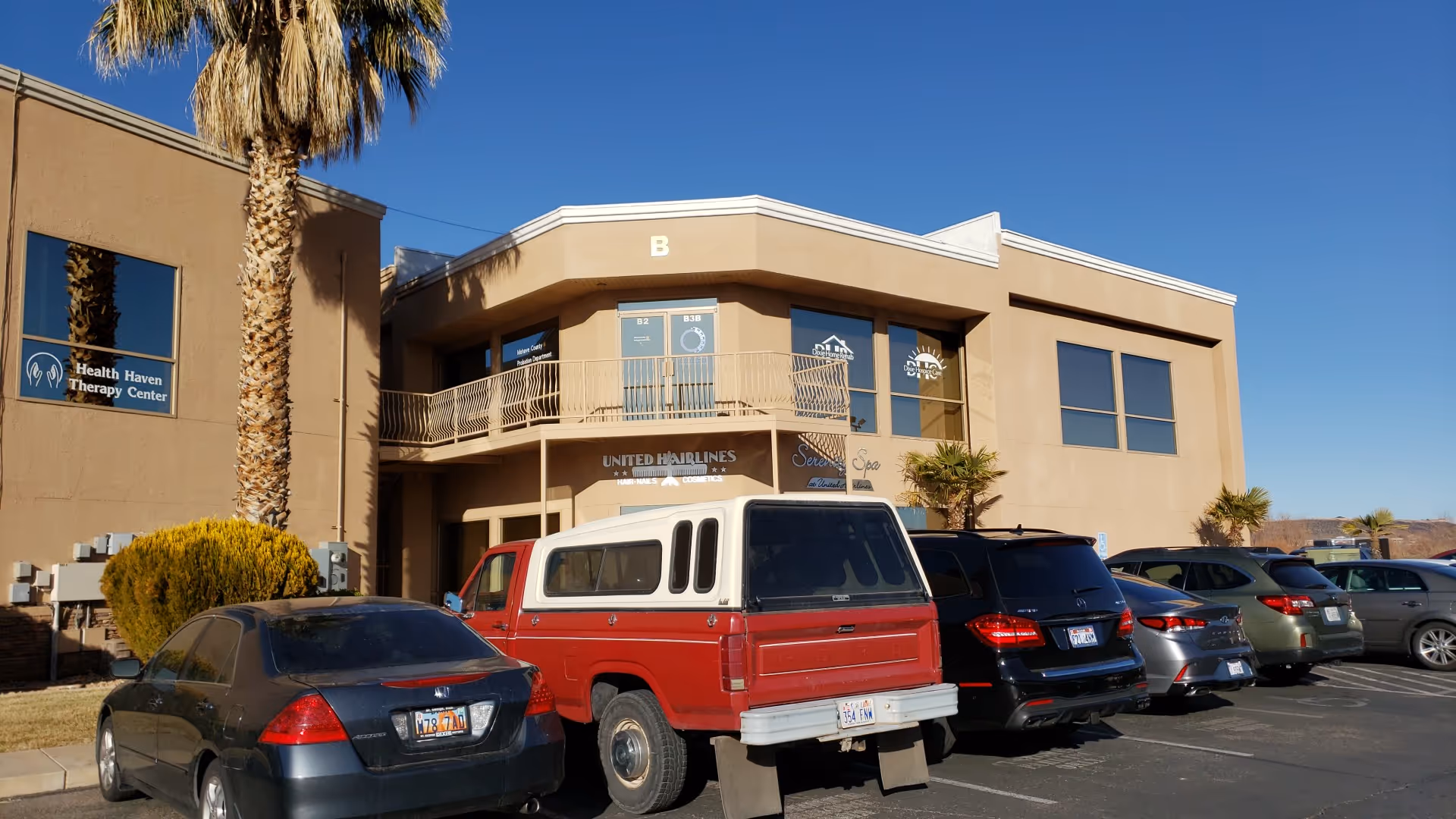 Exterior view of a tan building with a palm tree, parking lot, and blue sky.