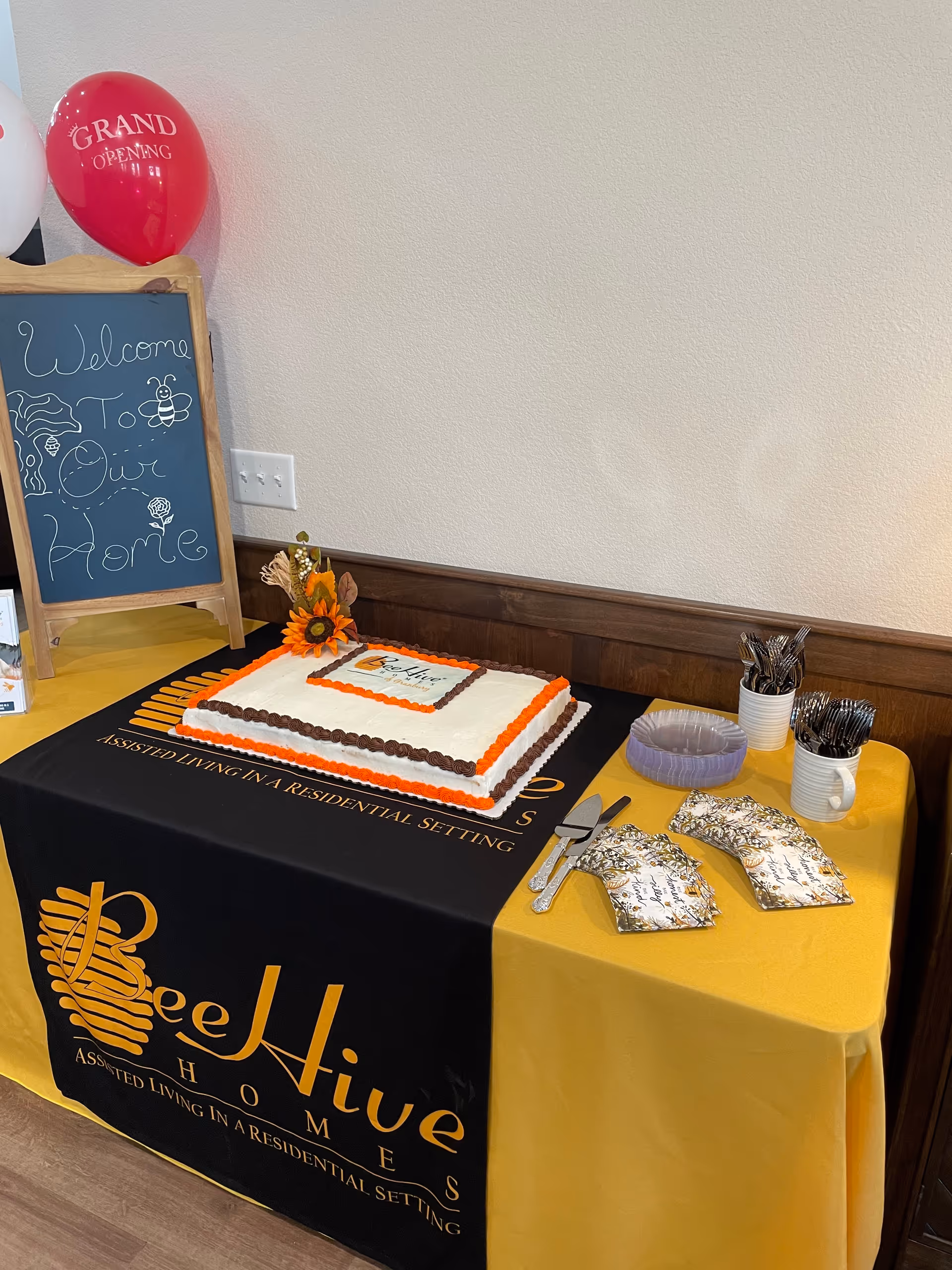 Indoor welcome display with cake on a BeeHive Homes table, chalkboard sign, and balloons.