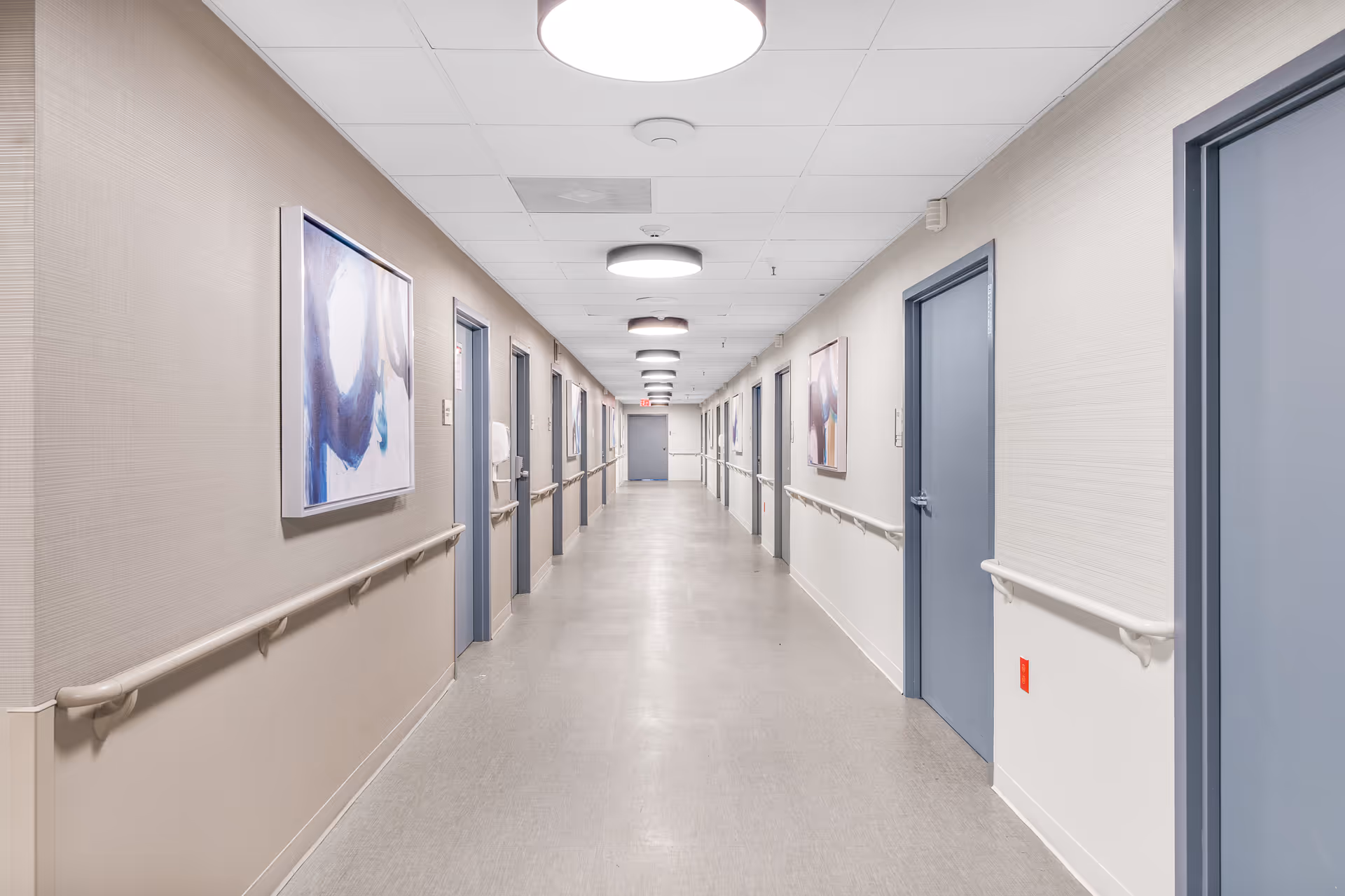 Long corridor in a senior living facility with blue doors, wall art, and handrails.