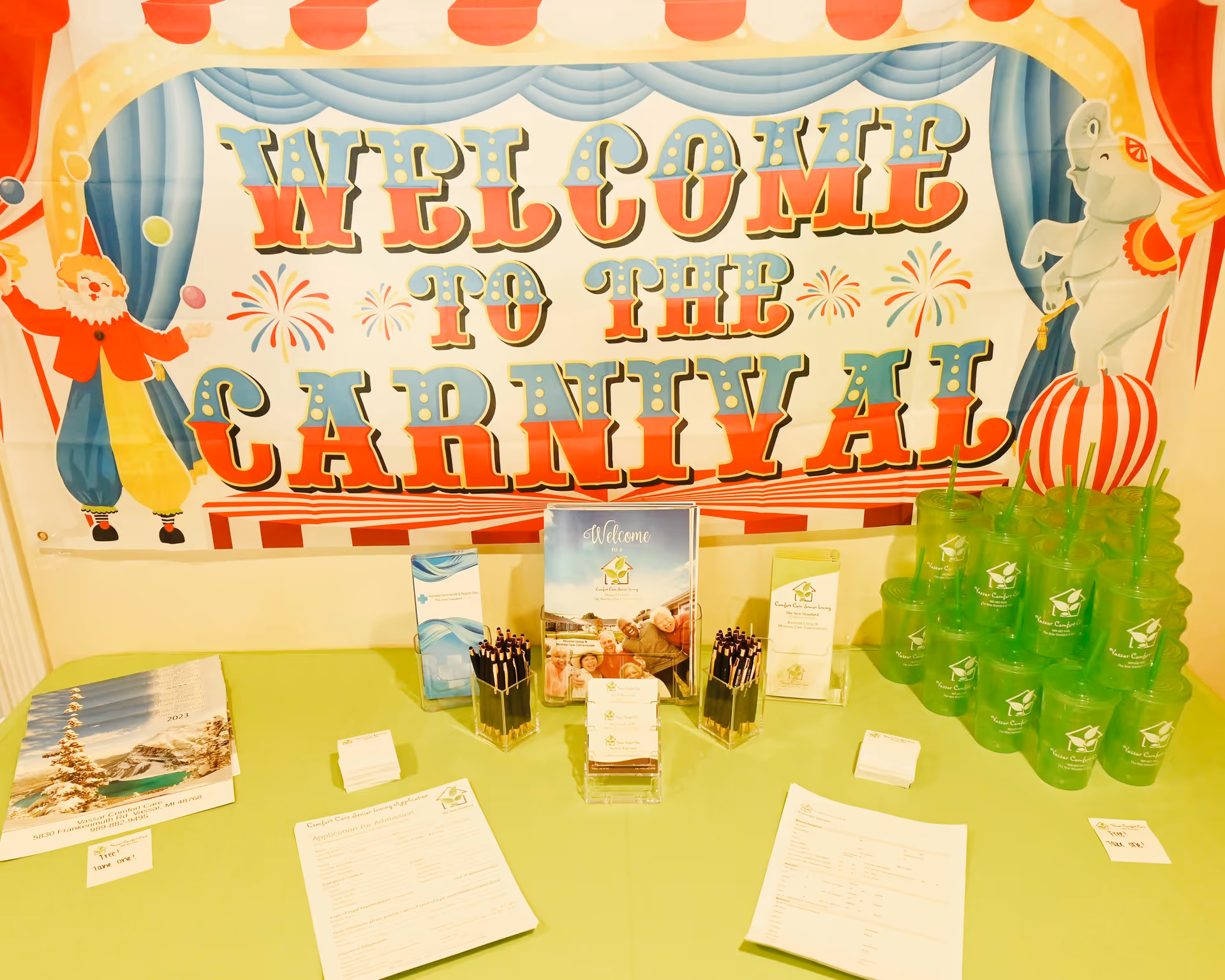 Bright carnival-themed welcome banner over a table with brochures and green cups indoors.