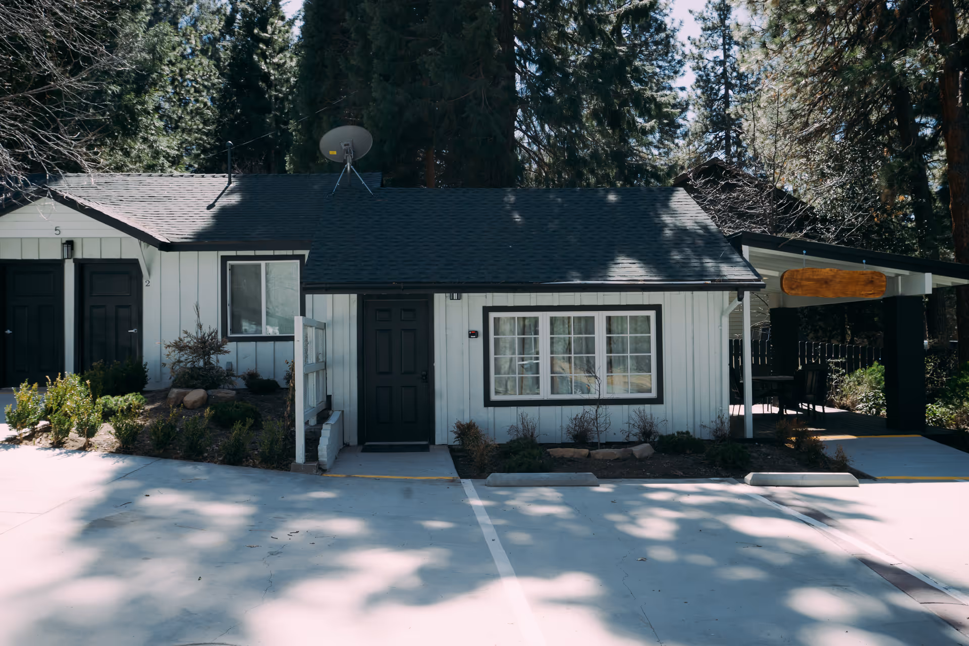 Single-story building exterior with a dark front door, window, and carport among trees.