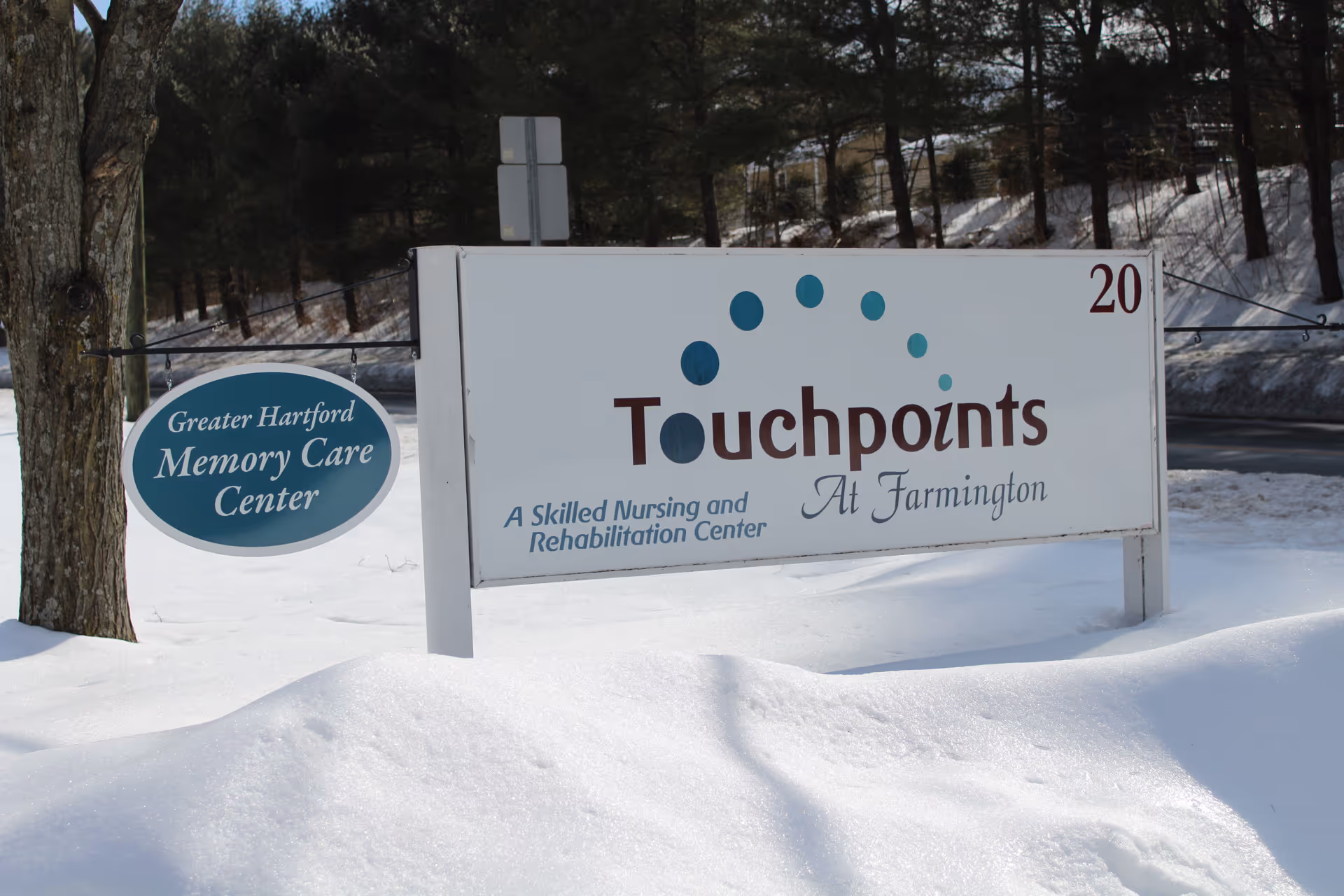 Outdoor sign for Touchpoints at Farmington, a skilled nursing and rehabilitation center, with an additional smaller sign for Greater Hartford Memory Care Center. The signs are surrounded by snow and trees in the background.