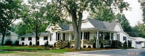 White, single-story building with a covered front porch, railing, and surrounding trees.