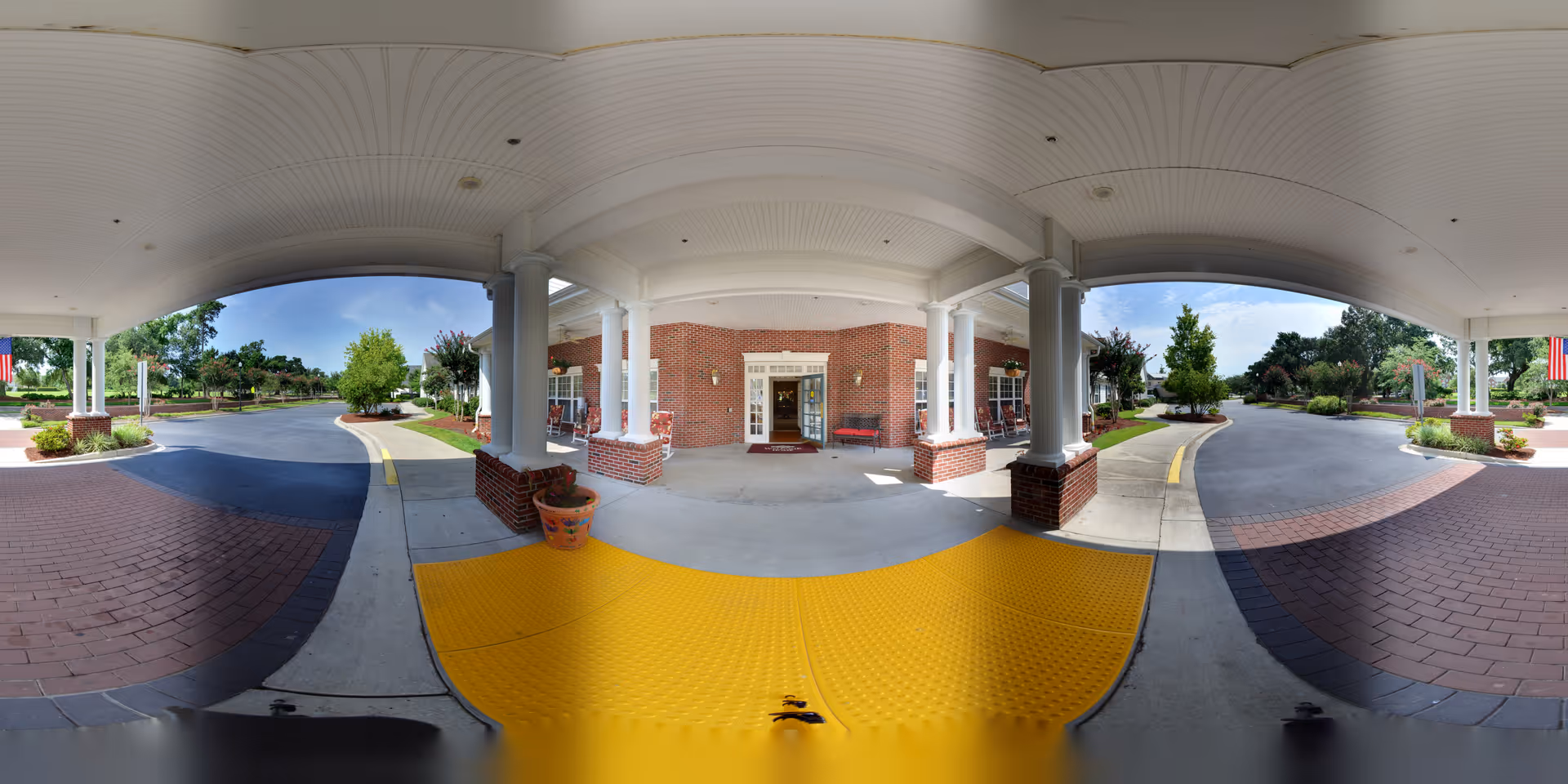 Covered entrance area of a senior living facility with white columns and brick walls. There is a yellow tactile paving strip on the ground for accessibility, potted plants, and a driveway leading up to the entrance. Trees and landscaping surround the driveway under a clear blue sky.